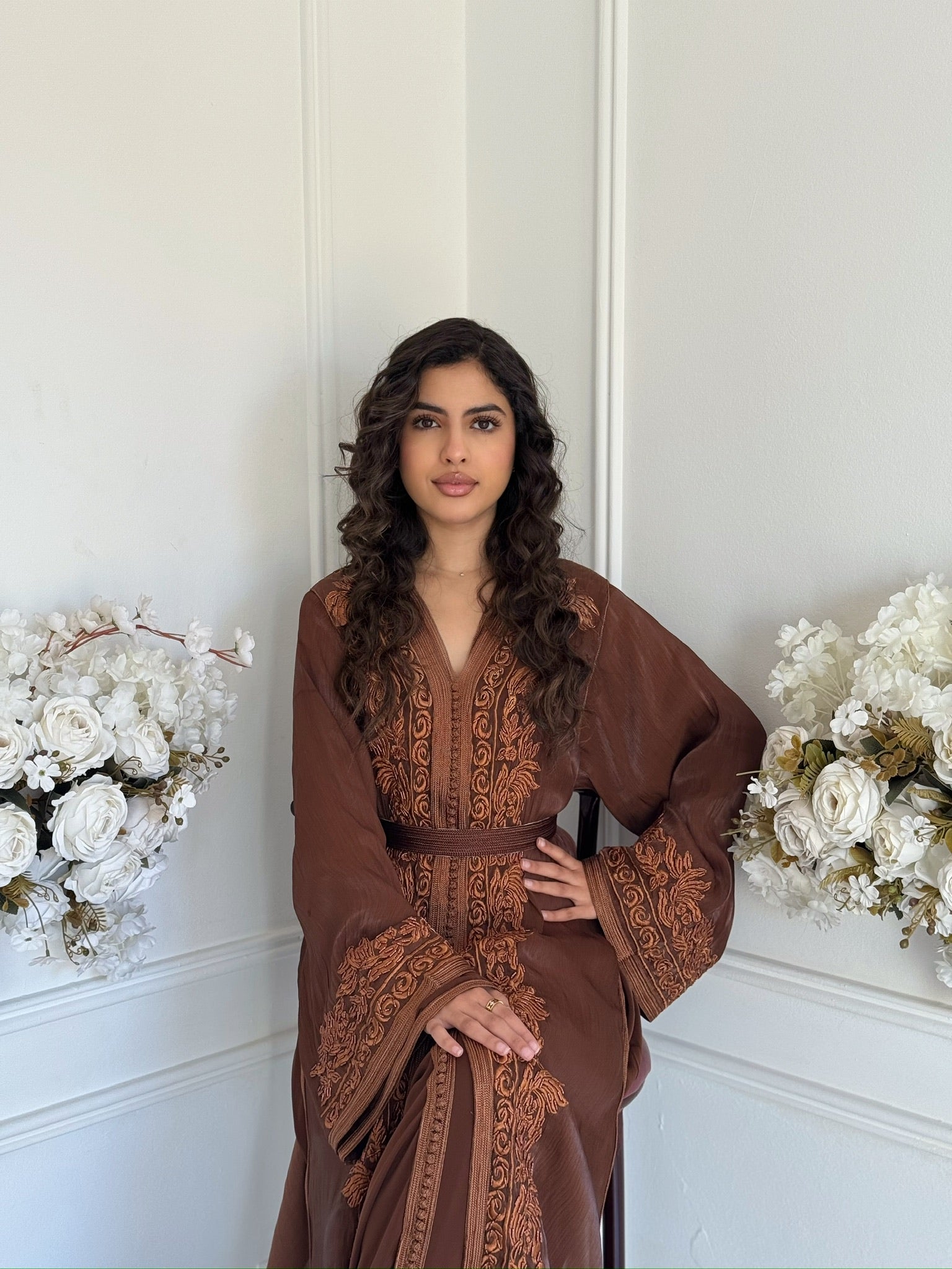 Brown Moroccan takchita with elegant embroidery displayed in a refined interior with white floral decor.