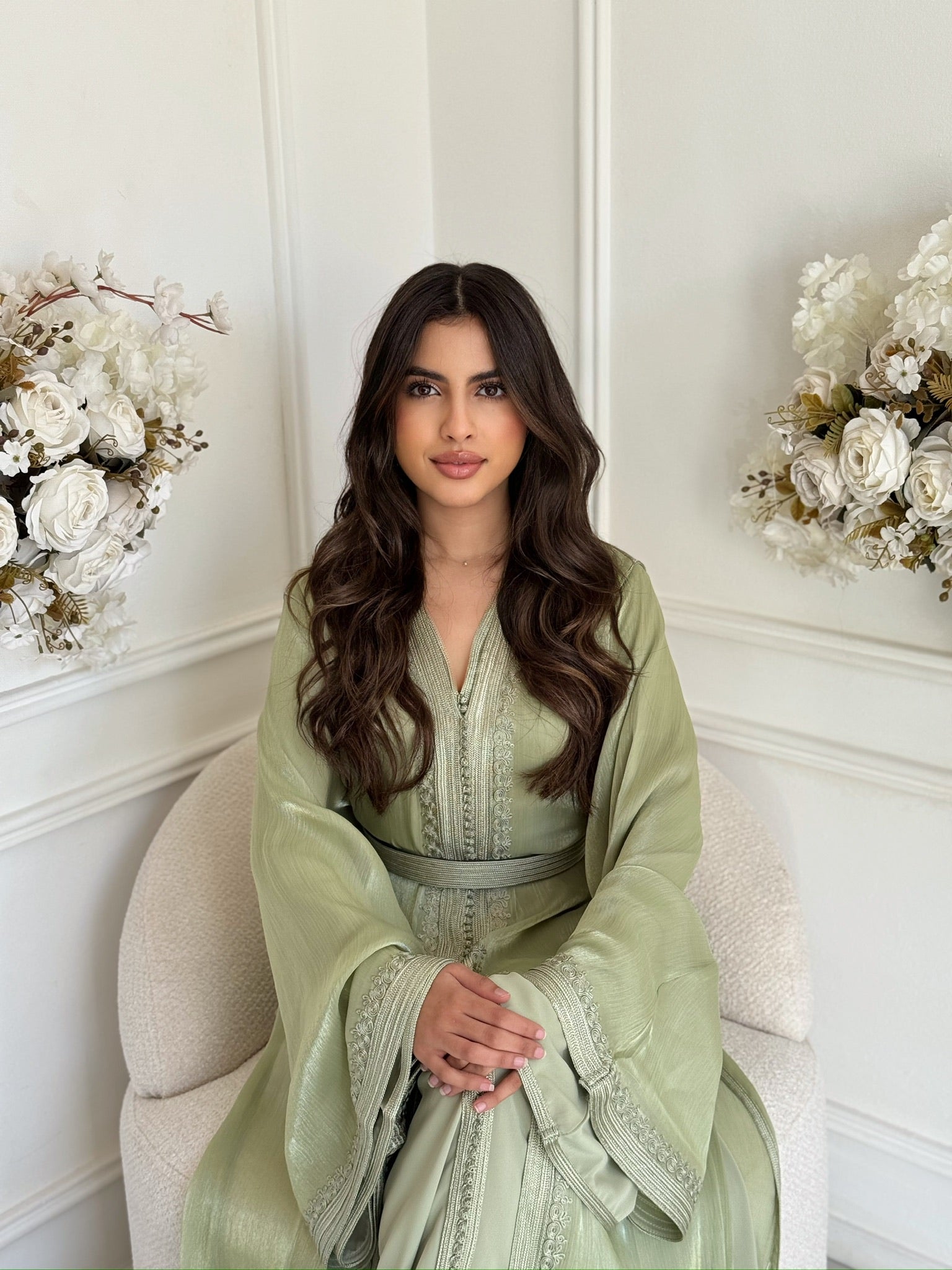 Model wearing a soft green Moroccan takchita sitting with floral décor in a light room